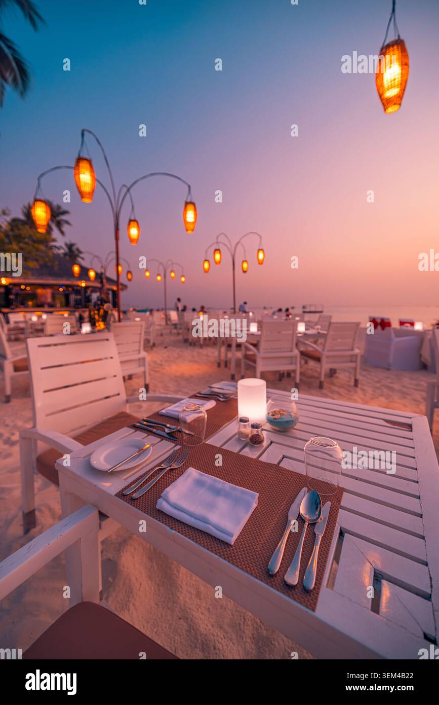 Lanterne sospese sopra il tavolo da pranzo sulla spiaggia al crepuscolo, vista verticale, apparecchi eleganti sulla riva sabbiosa, serata romantica e calda in un resort tropicale scenografico Foto Stock
