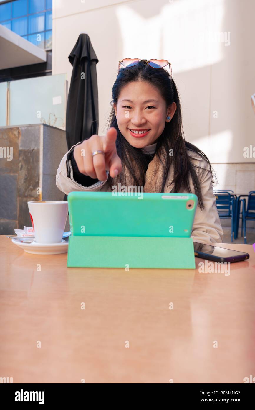 Felice donna asiatica che punta alla fotocamera durante la videochiamata al caffè all'aperto con borse Foto Stock