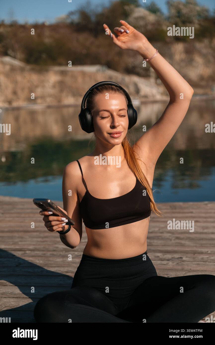 Donna attraente che ascolta playlist con le cuffie wireless. Bella donna che si rilassa accanto all'acqua durante il pomeriggio soleggiato. Concetto di equilibrio benessere. Foto Stock