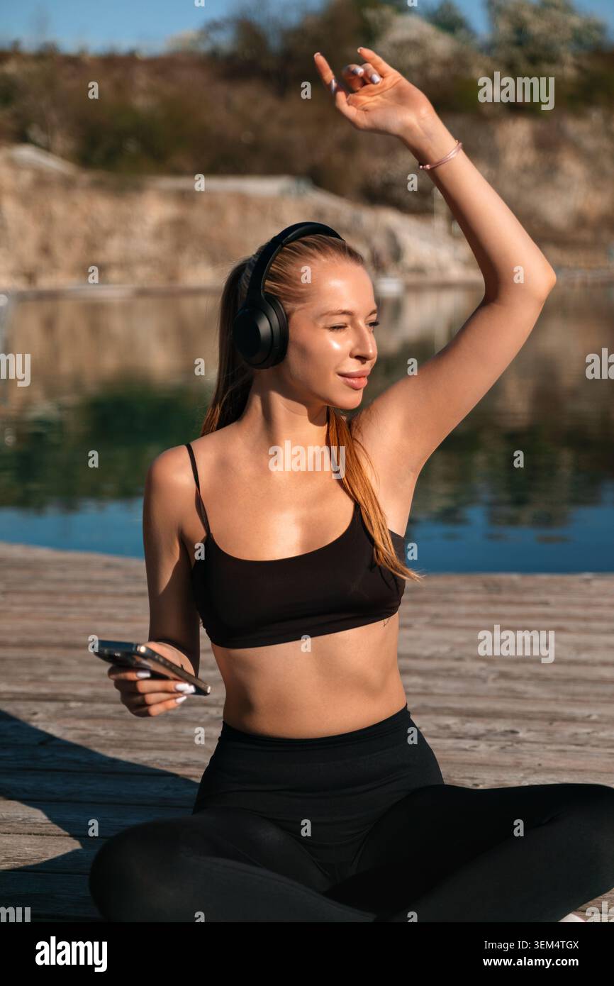 Una donna si siede accanto all'acqua, ascoltando musica e godendosi la routine di allenamento sotto il cielo azzurro. Foto Stock
