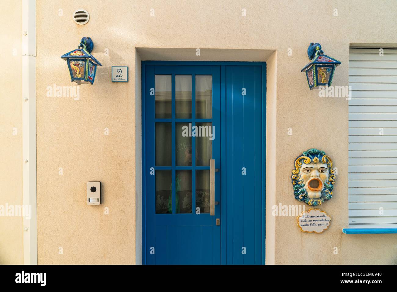 Vista di una porta in legno blu vibrante con pannelli di vetro, lanterne decorative e una maschera in ceramica su una parete beige a Marsaskala, regione meridionale, Malta. Foto Stock