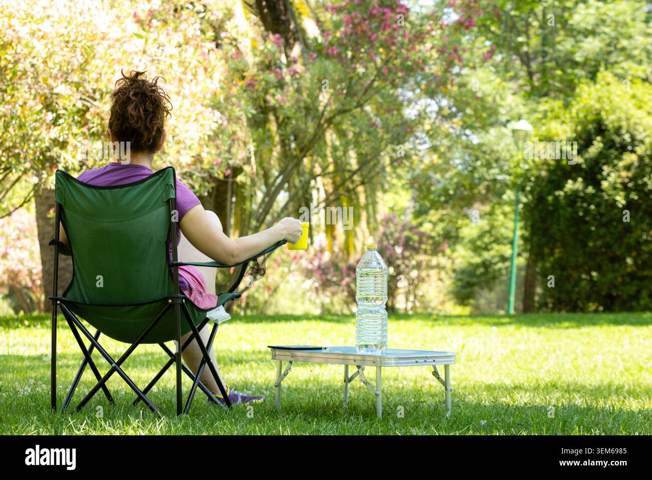 Una donna che si rilassa su una sedia da campeggio con un drink in un bicchiere giallo nel parco Foto Stock