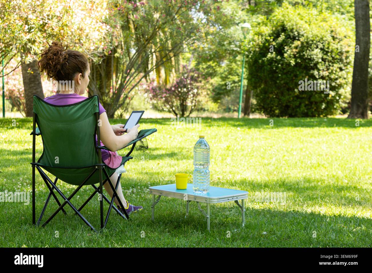 Una donna che legge su una sedia da campeggio con un drink e una bottiglia d'acqua sul tavolo, nel parco Foto Stock
