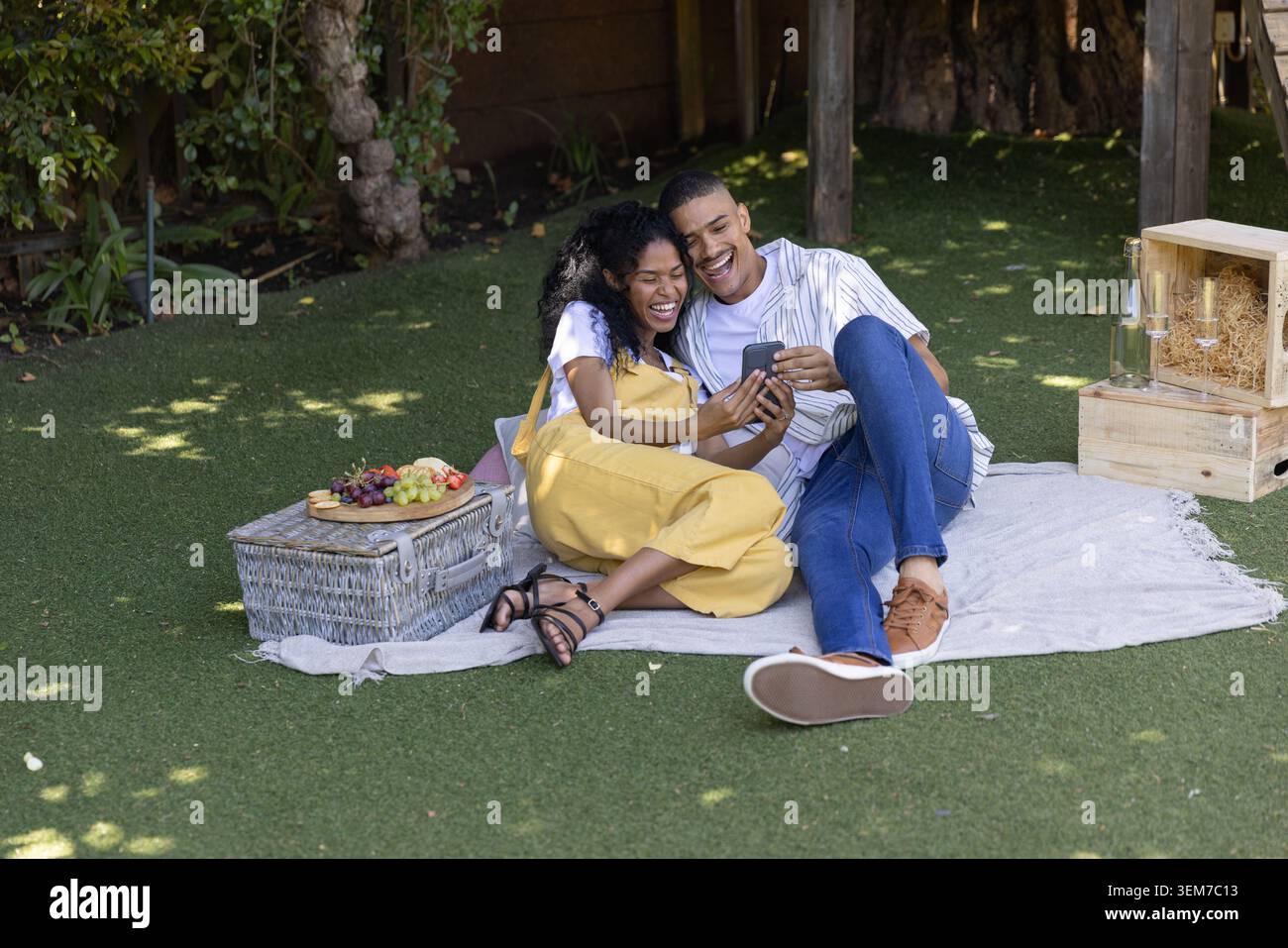 Coppia afroamericana che ride, che si piega insieme mentre guarda lo smartphone sulla coperta da picnic Foto Stock