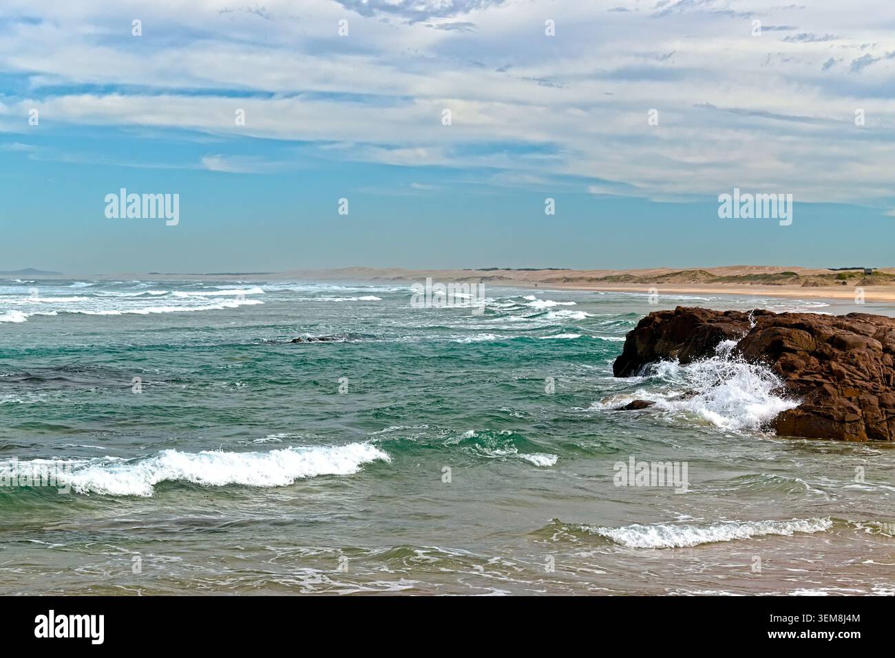Le onde turchesi e blu intenso si scontrano con una grande formazione rocciosa vicino alla costa, facendo oscillare schiuma bianca intorno ai suoi bordi robusti. Foto Stock