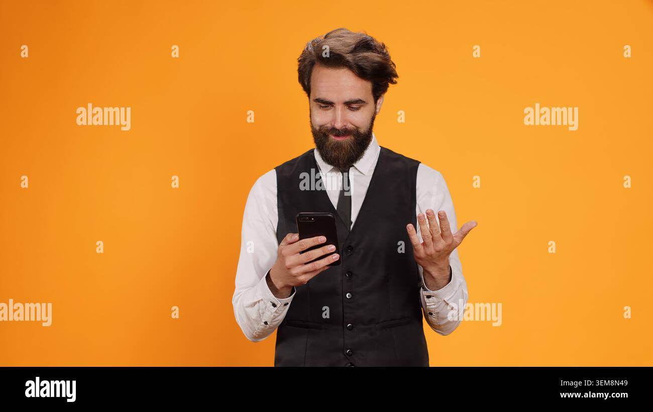 Cameriere professionista che controlla il telefono prima di servire i clienti al tavolo del ristorante. Elegante maggiordomo di lusso che lavora nel settore culinario, leggendo testi sull'app social media, servizio formale. Foto Stock
