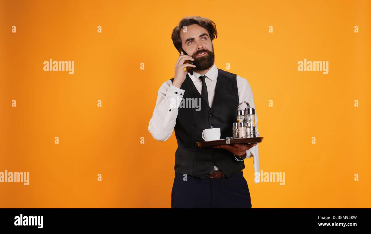 Cameriere del ristorante su chiamata tramite smartphone che effettua prenotazioni per cene eleganti, lavora nel settore della ristorazione su sfondo giallo. Uomo in abbigliamento formale che parla con i clienti su una linea telefonica remota. Foto Stock
