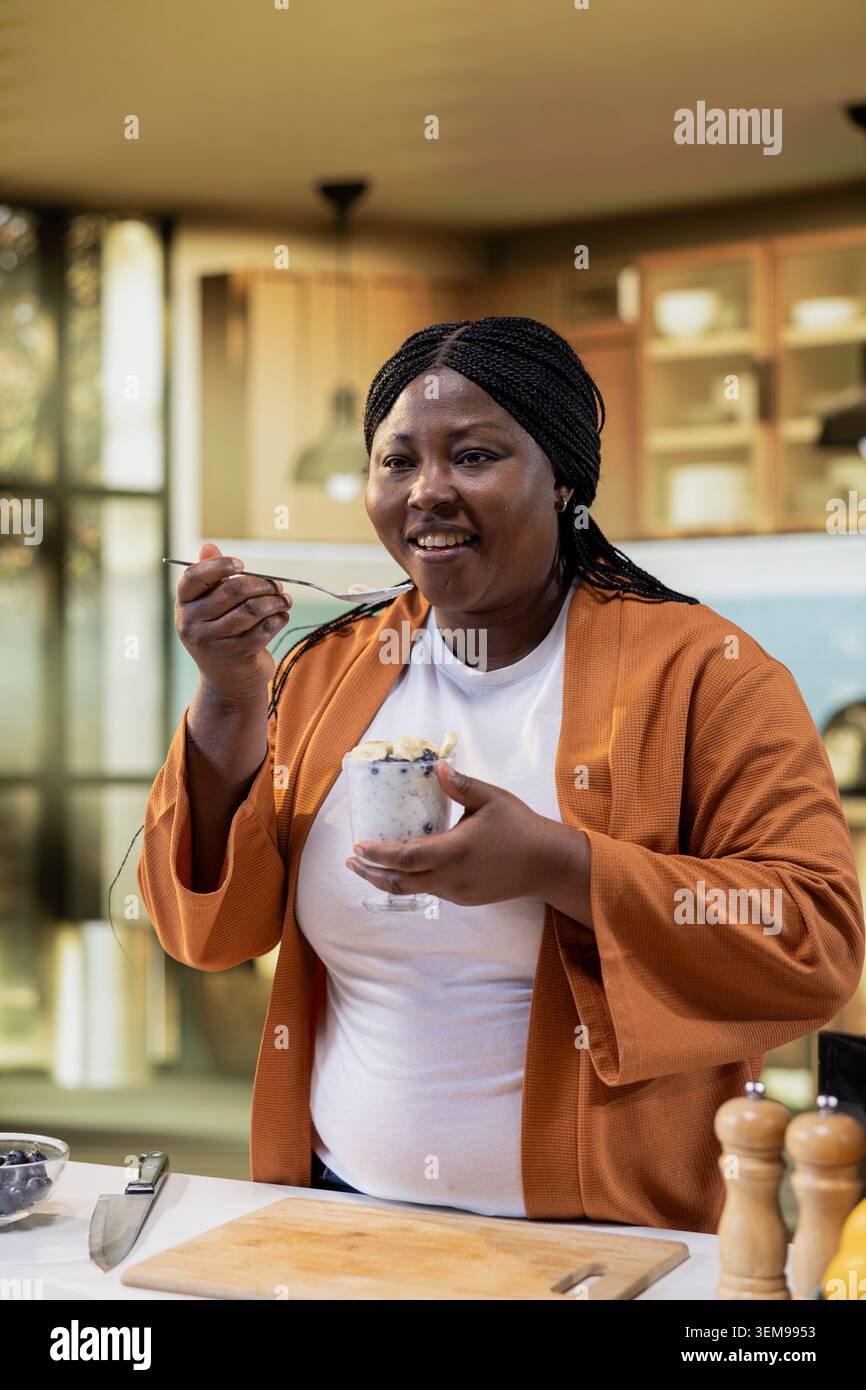 Chef afroamericano che fa un test del gusto con la ricetta della ciotola di chia al bancone. Influencer femminile che assaggia un budino di chia con yogurt e mirtilli, registrando la preparazione dei pasti per il canale online. Foto Stock