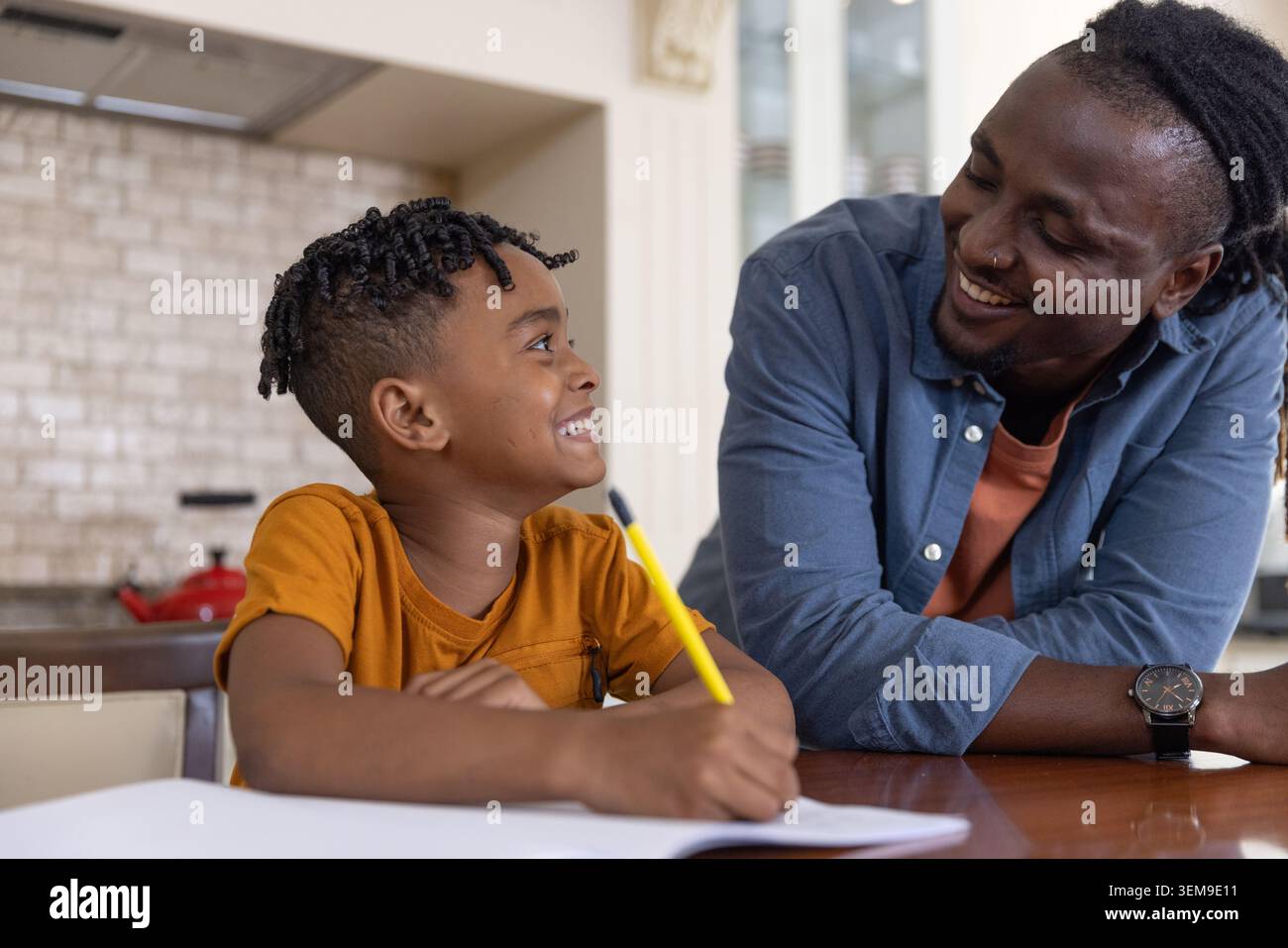 Papà afroamericano appoggiato sul tavolo, aiutando ragazzo a scrivere con matita gialla, notebook in cucina Foto Stock