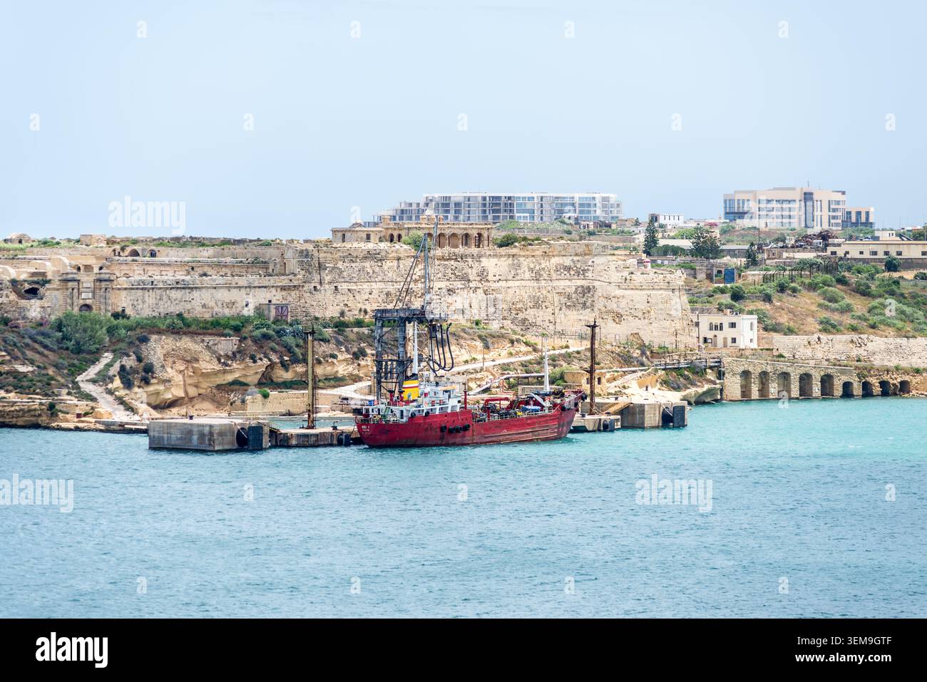 Kalkara, Malta - 16 maggio 2025: Nave da carico ormeggiata nella baia di Rinella vicino allo storico forte Ricasoli con fortificazioni costiere e turchese Mediterranea Foto Stock