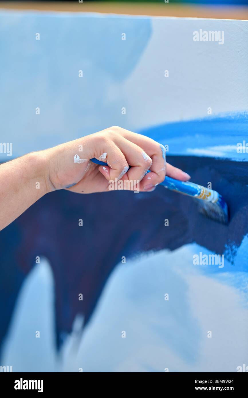 Pennello per mano dell'artista che aggiunge un colore blu intenso sulla tela durante il processo creativo di pittura. La luce naturale soffusa mette in risalto i tratti di pennello e la fusione Foto Stock