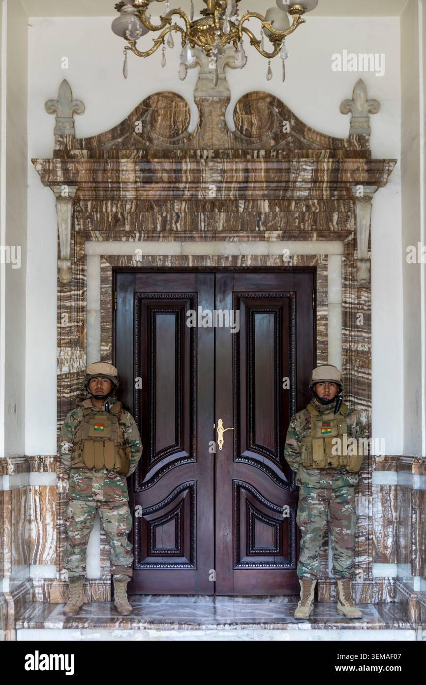 Due soldati boliviani armati con equipaggiamento da combattimento completo sono all'attenzione di fronte al grande ingresso di quello che sembra essere il Palacio de Gobierno o un cartello Foto Stock