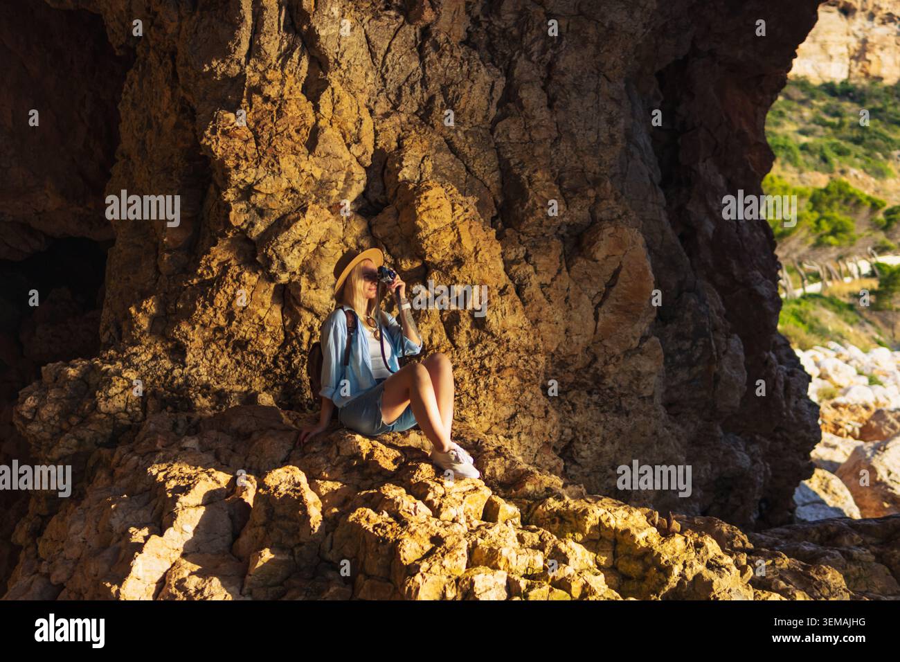 Donna siede su una roccia all'interno di una grotta costiera, fotografando il paesaggio panoramico mentre la luce del sole filtra, godendosi un viaggio avventuroso, esplorando la natura Foto Stock