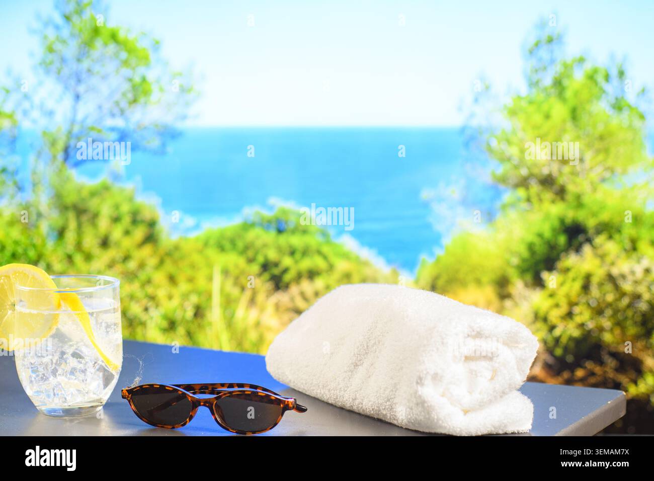 Acqua fredda al limone con ghiaccio, occhiali da sole e asciugamano bianco su una terrazza soleggiata affacciata sul Mar Mediterraneo e sulla vegetazione costiera. Foto Stock
