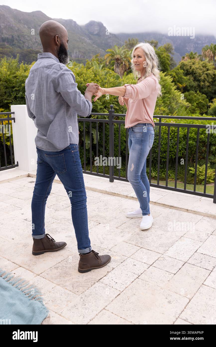 Diverse coppie senior che ballano e tengono le mani sulla terrazza piastrellata vicino alla ringhiera nera e alla coperta piegata Foto Stock