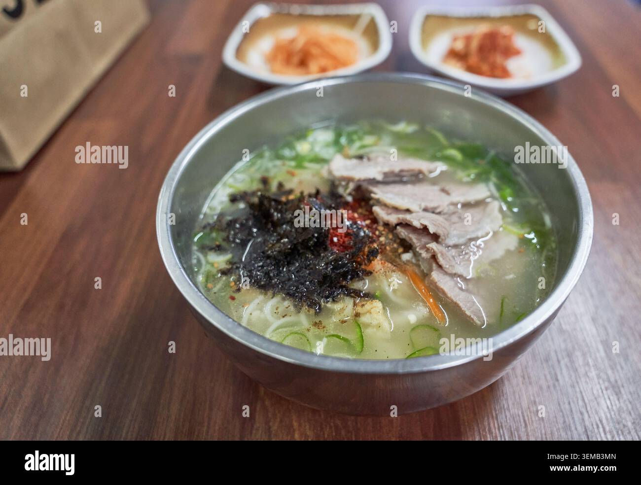 Piatto tradizionale dell'isola di Jeju con filamenti di grano in ricco brodo osseo, condito con carne di maiale bollita a fette, alghe marine e spezie in un bacino metallico rotondo. Foto Stock