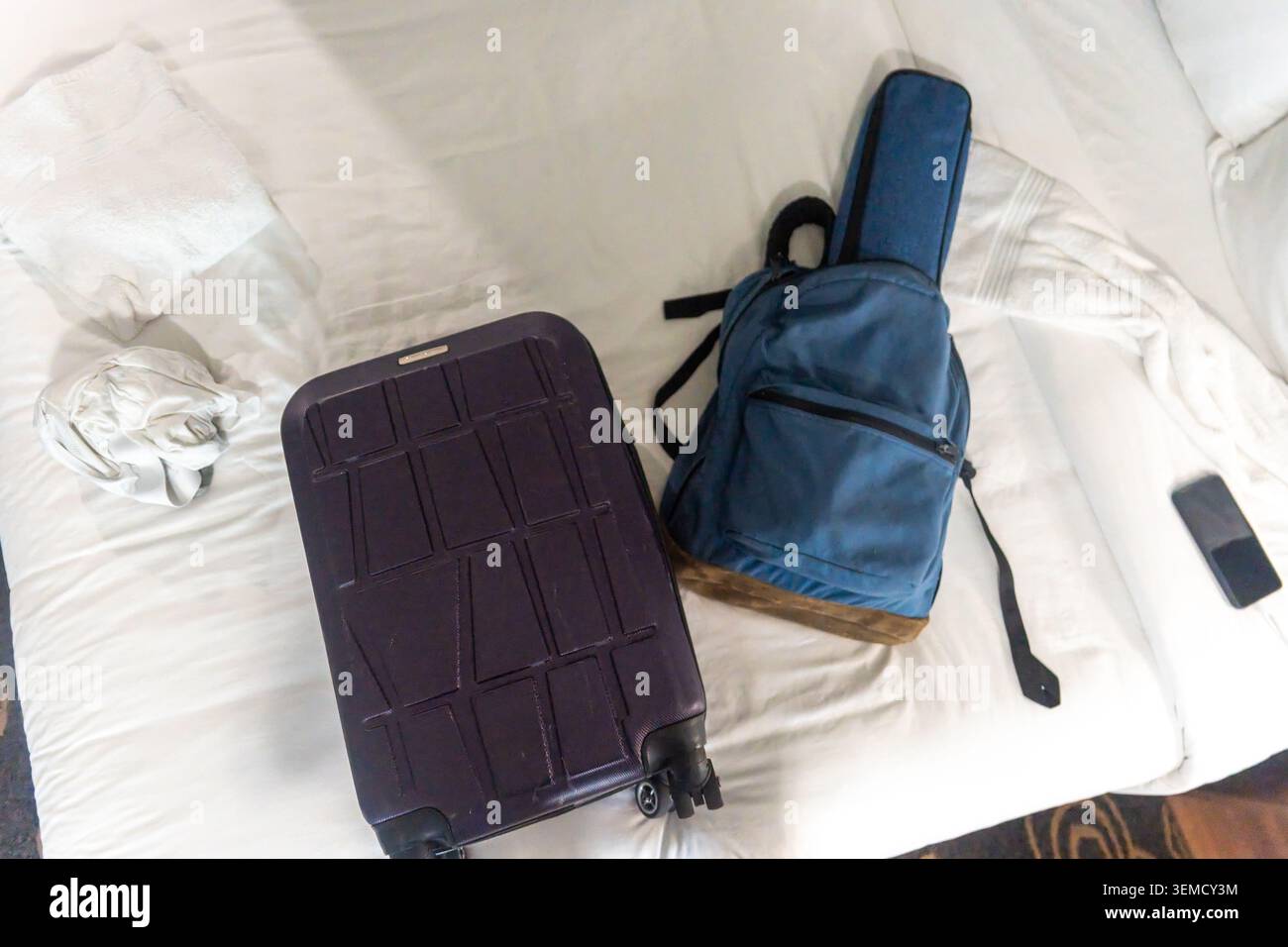 Una valigia rigida viola con ruote si trova su un letto bianco accanto a uno zaino blu. Uno smartphone si trova nelle vicinanze sul materasso, con cuscini e un cr Foto Stock