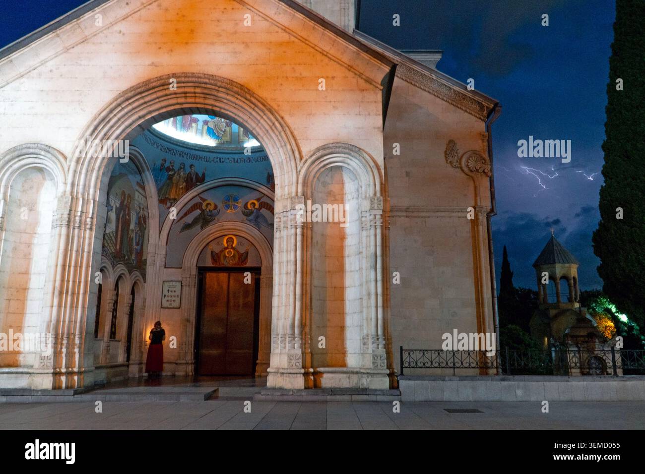 Tbilisi, Georgia, 6 giugno 2026. Una donna accende una candela mentre prega nella chiesa di San Giorgio di Kashueti mentre un fulmine colpisce il cielo di Tbilisi, Georgi Foto Stock