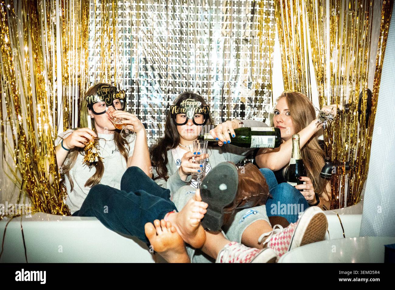 Tre giovani donne siedono in una vasca da bagno circondata da rastrellatori dorati, tenendo bottiglie di champagne e bicchieri, condividendo un umore di festa giocoso in un indo festivo Foto Stock