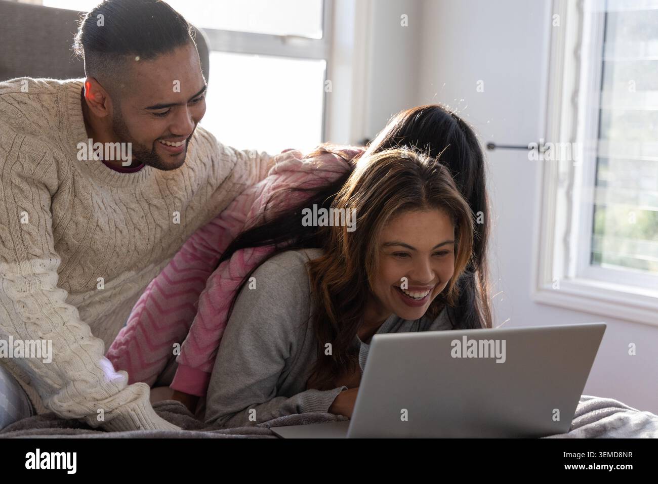 Famiglia di tre persone sdraiata sul letto, papà in maglione crema, bambino in felpa con cappuccio rosa che guarda il laptop Foto Stock
