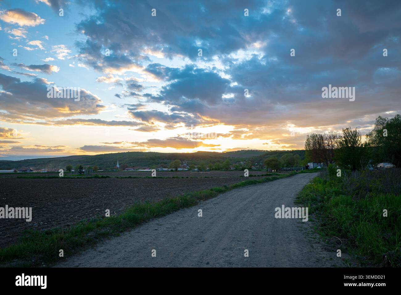 Tramonto su strade sterrate rurali e terreni agricoli con orizzonte del villaggio e suggestivo paesaggio nuvoloso Foto Stock