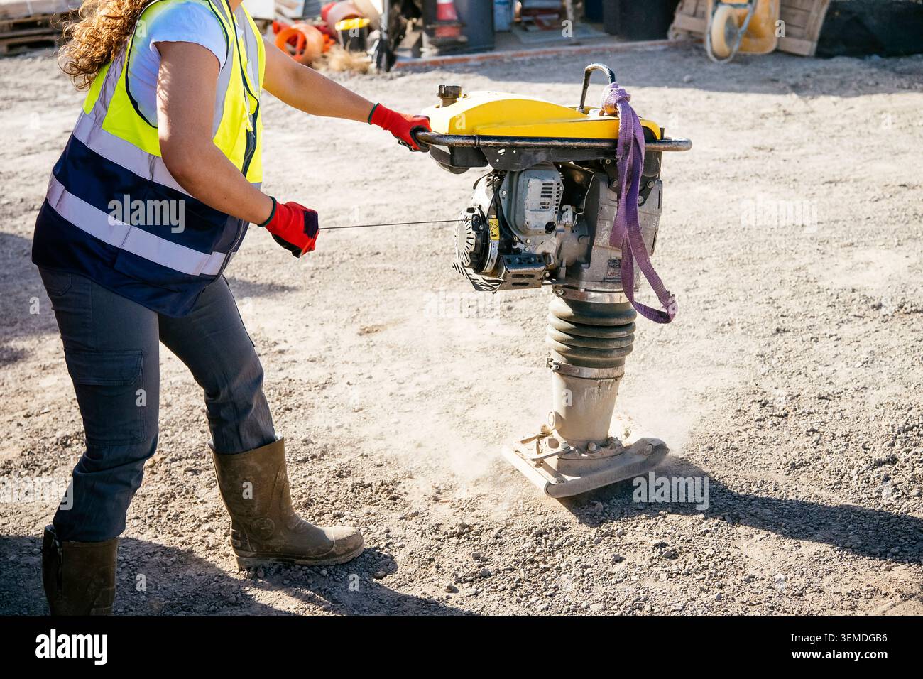 Una lavoratrice edile che indossa giubbotti di sicurezza e guanti aziona un compattatore vibrante su un cantiere di ghiaia, livellando il terreno su strada o su terreni accidentati Foto Stock