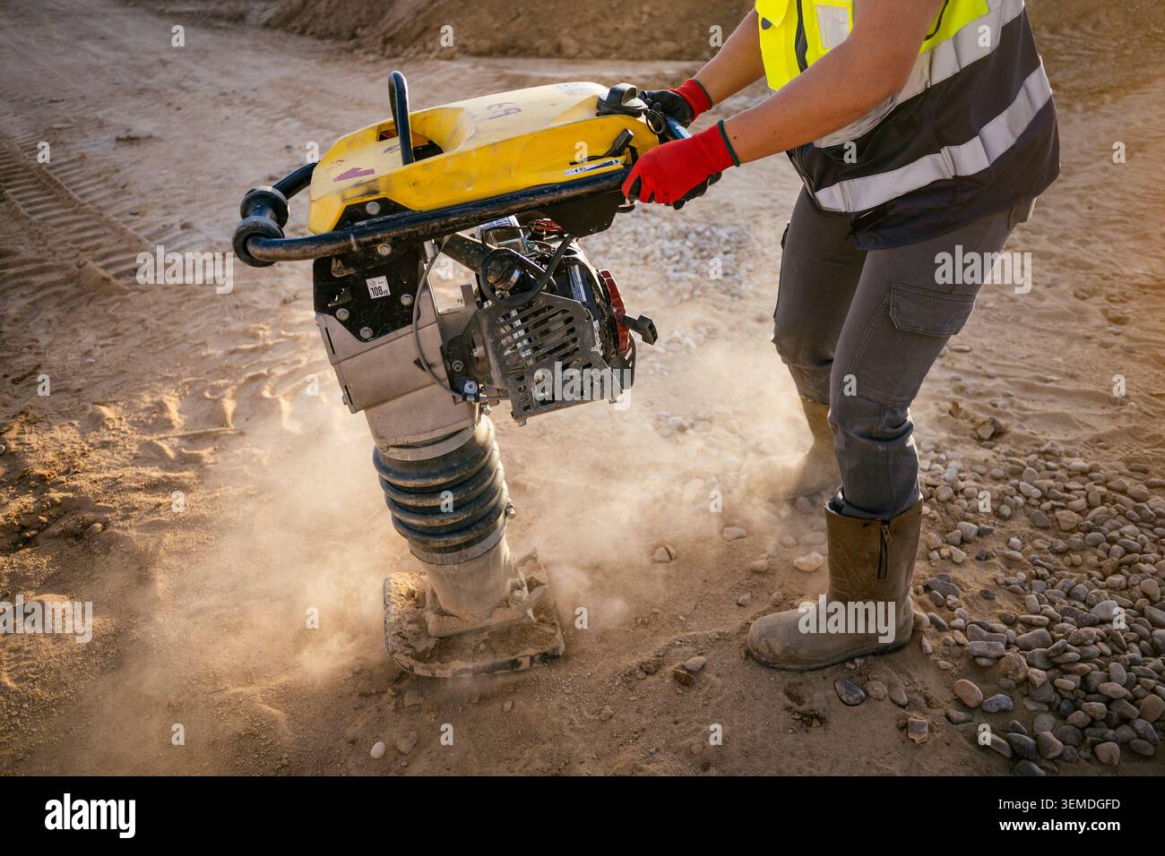 Una lavoratrice edile in giubbotto di sicurezza e guanti utilizza un compattatore vibrante per compattare il terreno e la ghiaia in un cantiere stradale polveroso di giorno Foto Stock
