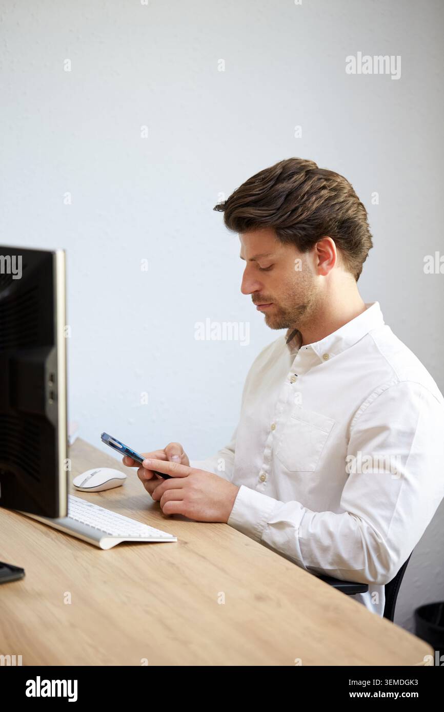 Un giovane professionista con una camicia bianca si concentra con attenzione sul suo smartphone in una moderna scrivania da ufficio. Sono visibili un computer e un mouse, evidenziando un Foto Stock