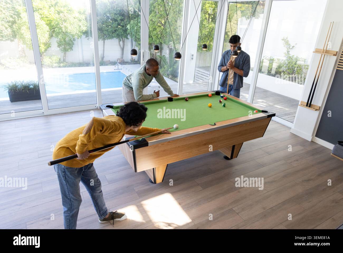 Diversi amici maschi che giocano su un tavolo in feltro verde tenendo spunti in una moderna sala giochi vicino a porte di vetro Foto Stock