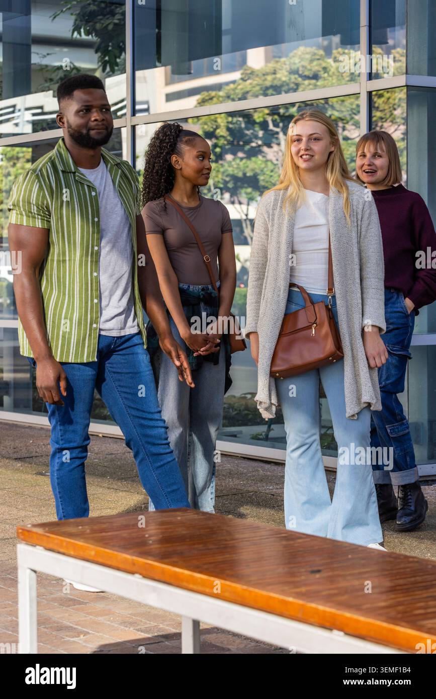 Gruppo di quattro amici che camminano e chiacchierano sul passaggio pavimentato da finestre di vetro e panchina di legno Foto Stock
