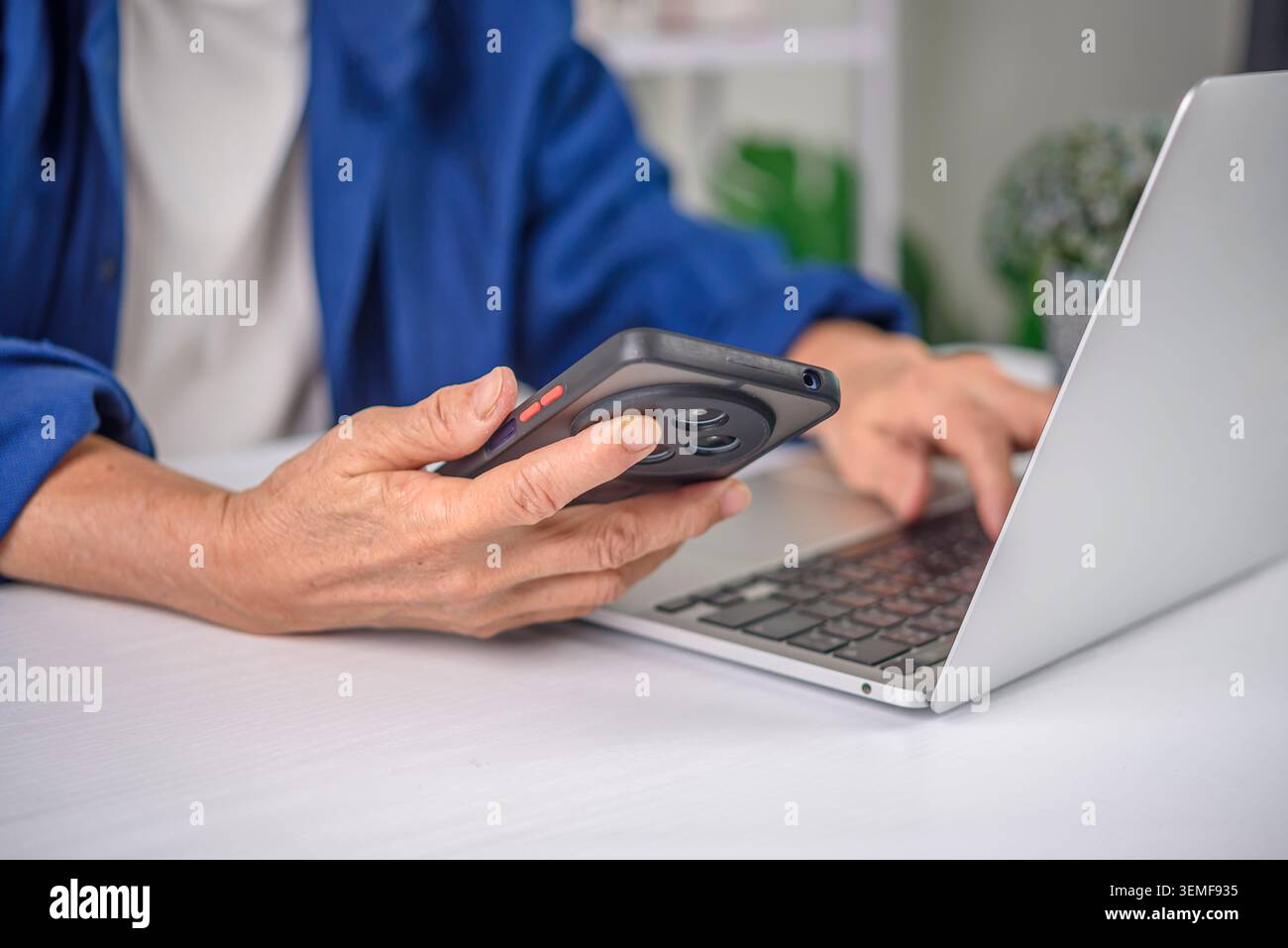Primo piano di una donna anziana che tiene le mani sullo smartphone e digita sulla tastiera del laptop. Donna d'affari anziana che lavora in multitasking con tecnologia, immissione dati e d Foto Stock