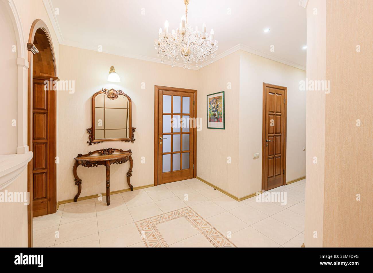 elegante corridoio con pareti color crema, pavimenti piastrellati, porte e finiture in legno, un lampadario di cristallo, una nicchia decorativa e un tavolo da toilette a specchio. Foto Stock
