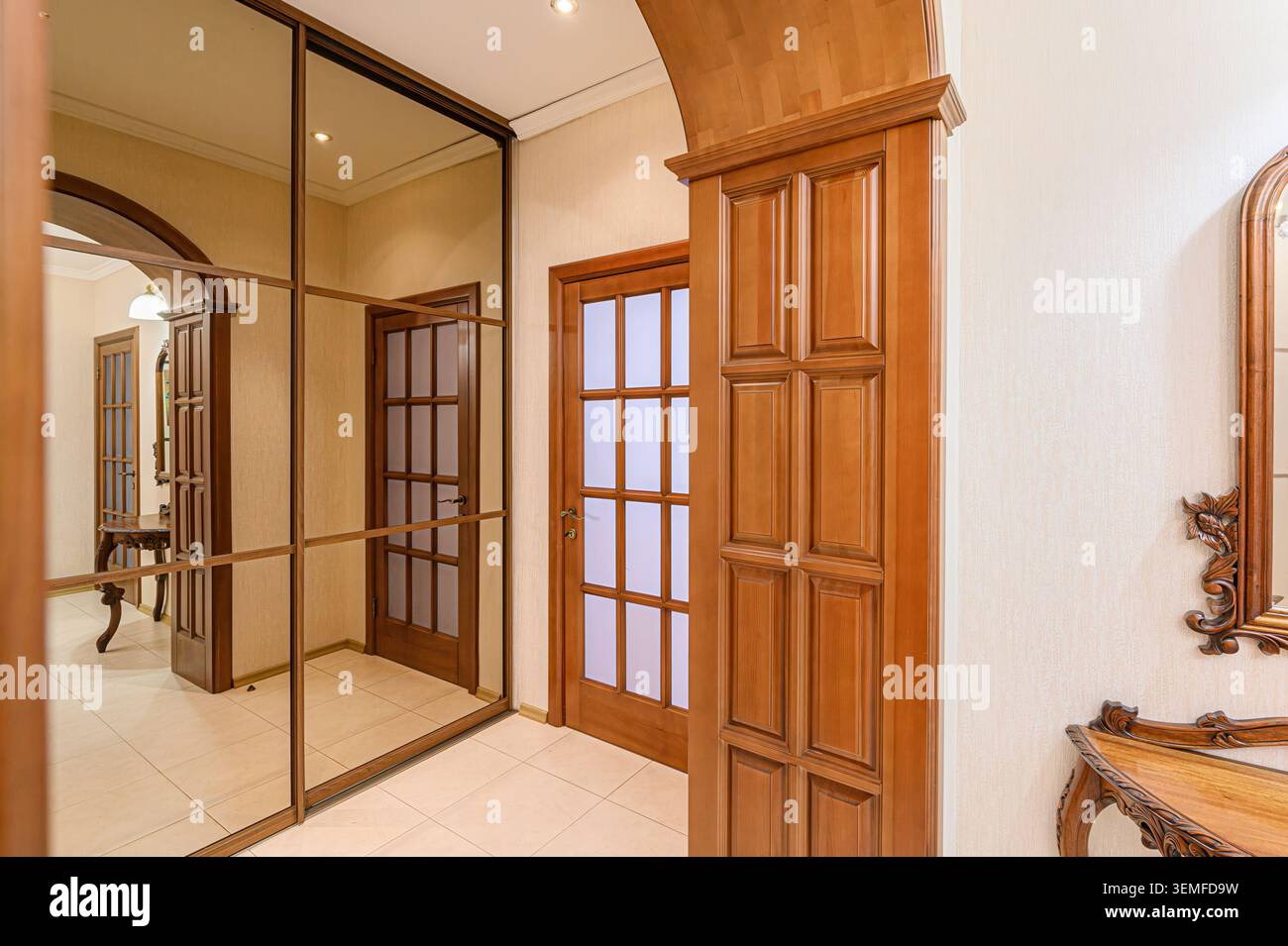 elegante corridoio con pareti color crema, pavimenti piastrellati, porte e finiture in legno, un lampadario di cristallo, una nicchia decorativa e un tavolo da toilette a specchio. Foto Stock