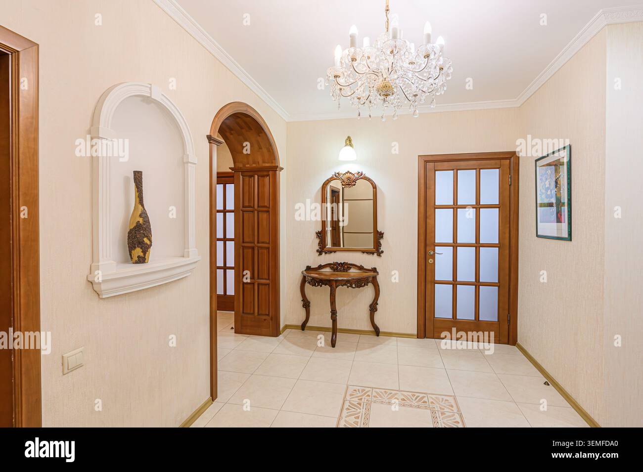 elegante corridoio con pareti color crema, pavimenti piastrellati, porte e finiture in legno, un lampadario di cristallo, una nicchia decorativa e un tavolo da toilette a specchio. Foto Stock