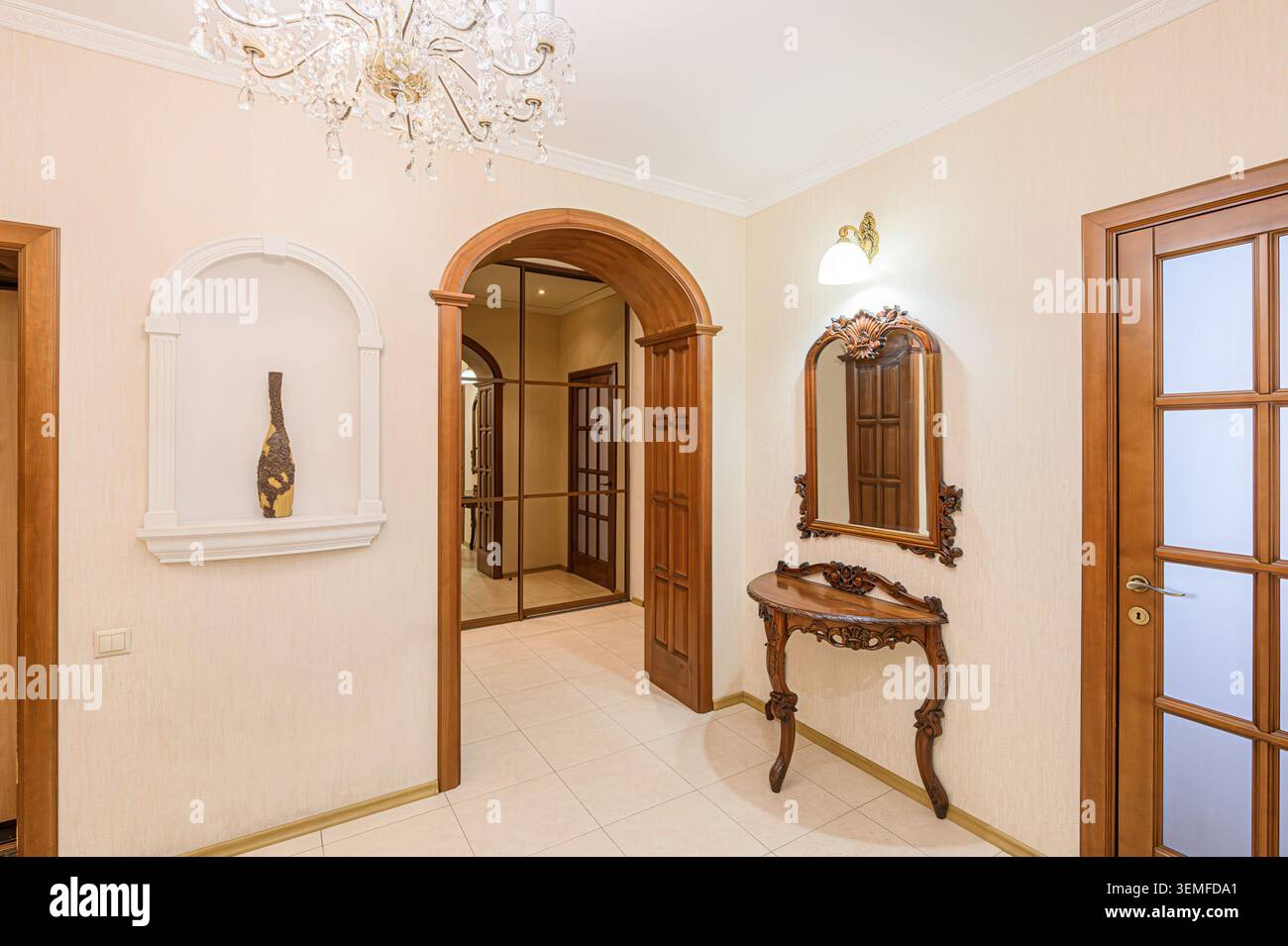 elegante corridoio con pareti color crema, pavimenti piastrellati, porte e finiture in legno, un lampadario di cristallo, una nicchia decorativa e un tavolo da toilette a specchio. Foto Stock