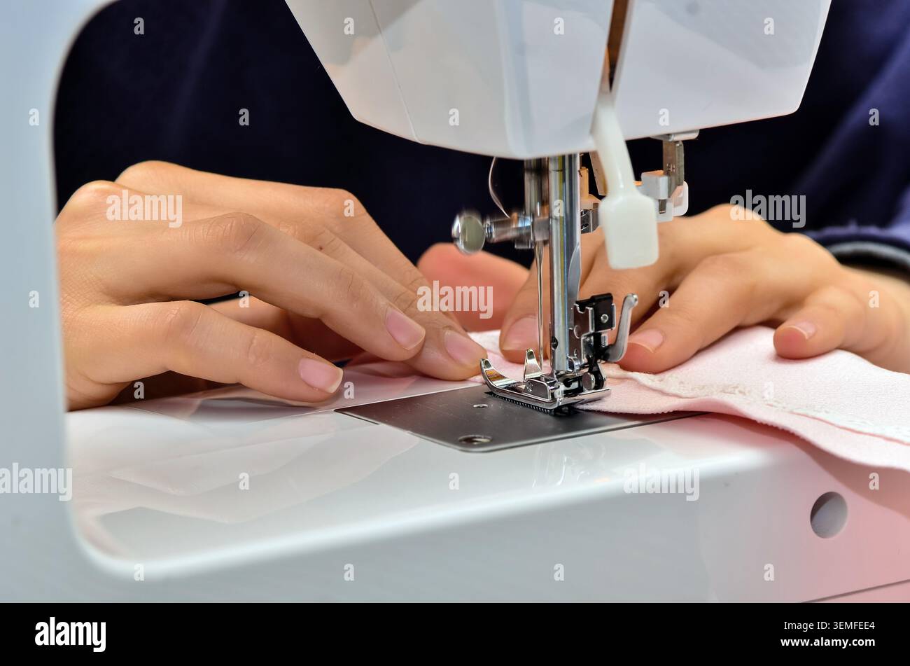 Primo piano di mani che guidano il tessuto rosa sotto il piede della pressa di una macchina da cucire domestica, cuciture e concetto di artigianato tessile. Foto Stock