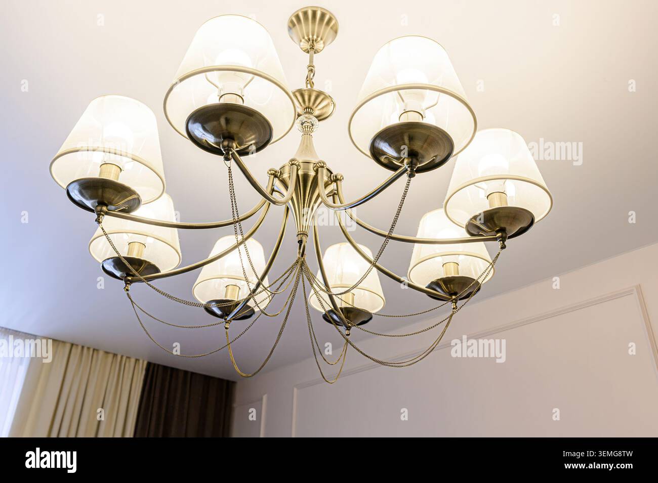 la foto del soffitto mostra un lampadario con otto lampade, ciascuna con un paralume bianco. È appeso a un soffitto bianco con modanatura e vista parziale delle tende. Foto Stock