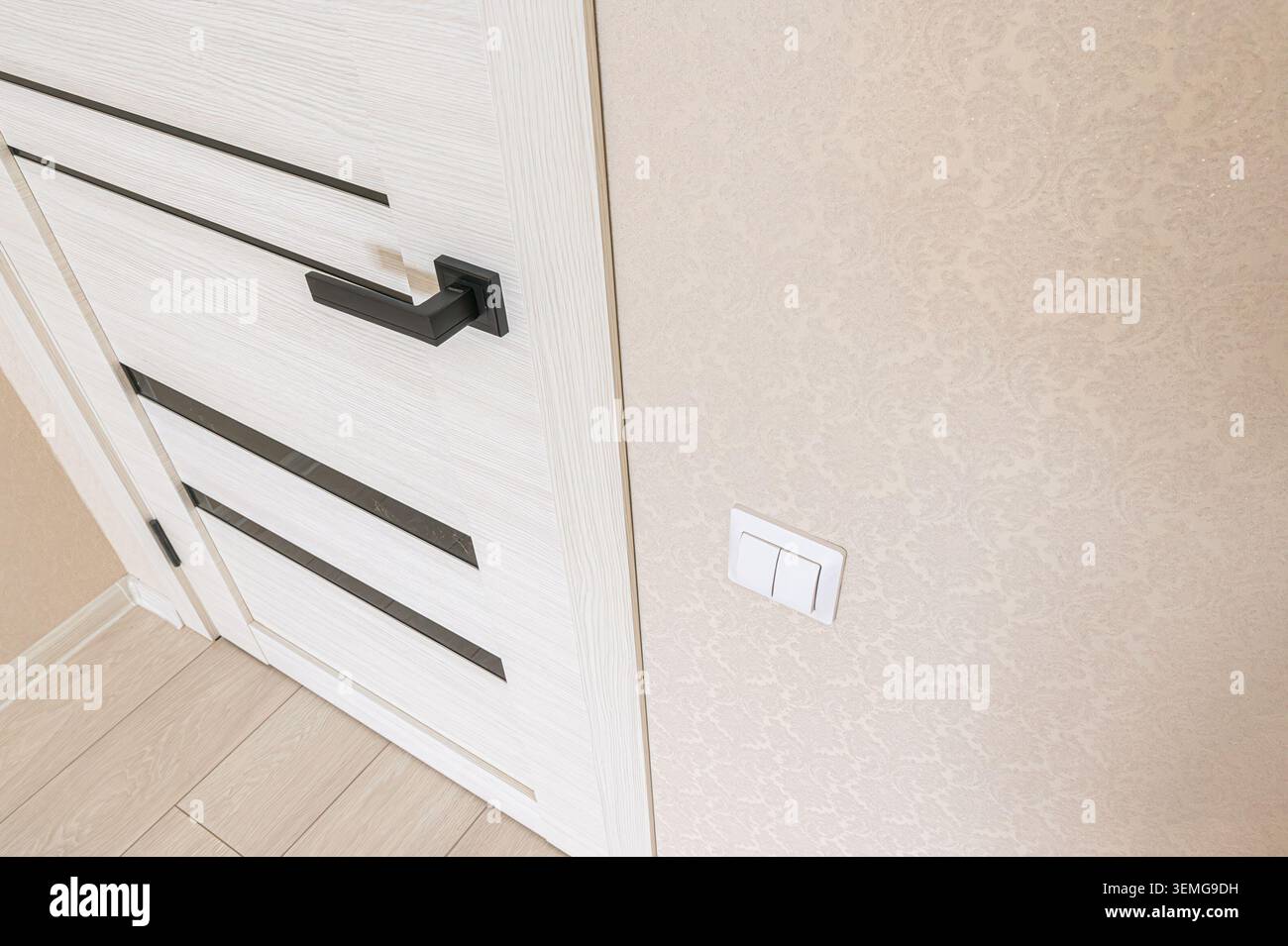 il primo piano presenta una moderna porta bianca con dettagli neri, una maniglia nera, un interruttore luce e una parete beige testurizzata. Il pavimento è di legno chiaro. Foto Stock