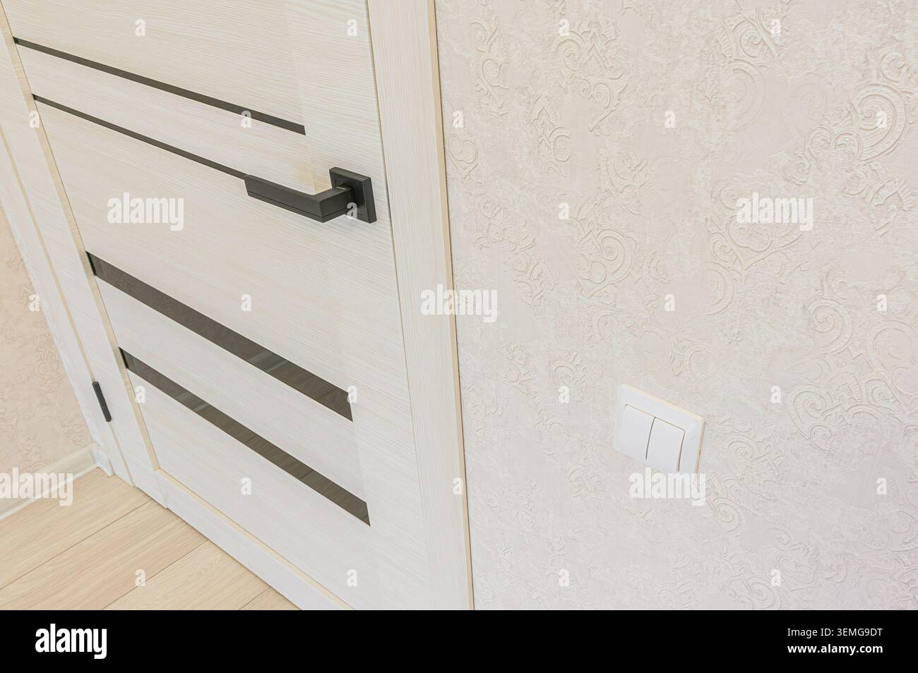 Foto degli interni con una moderna porta in legno chiaro con dettagli neri accanto a una parete beige con motivi geometrici e un interruttore luce bianco Foto Stock