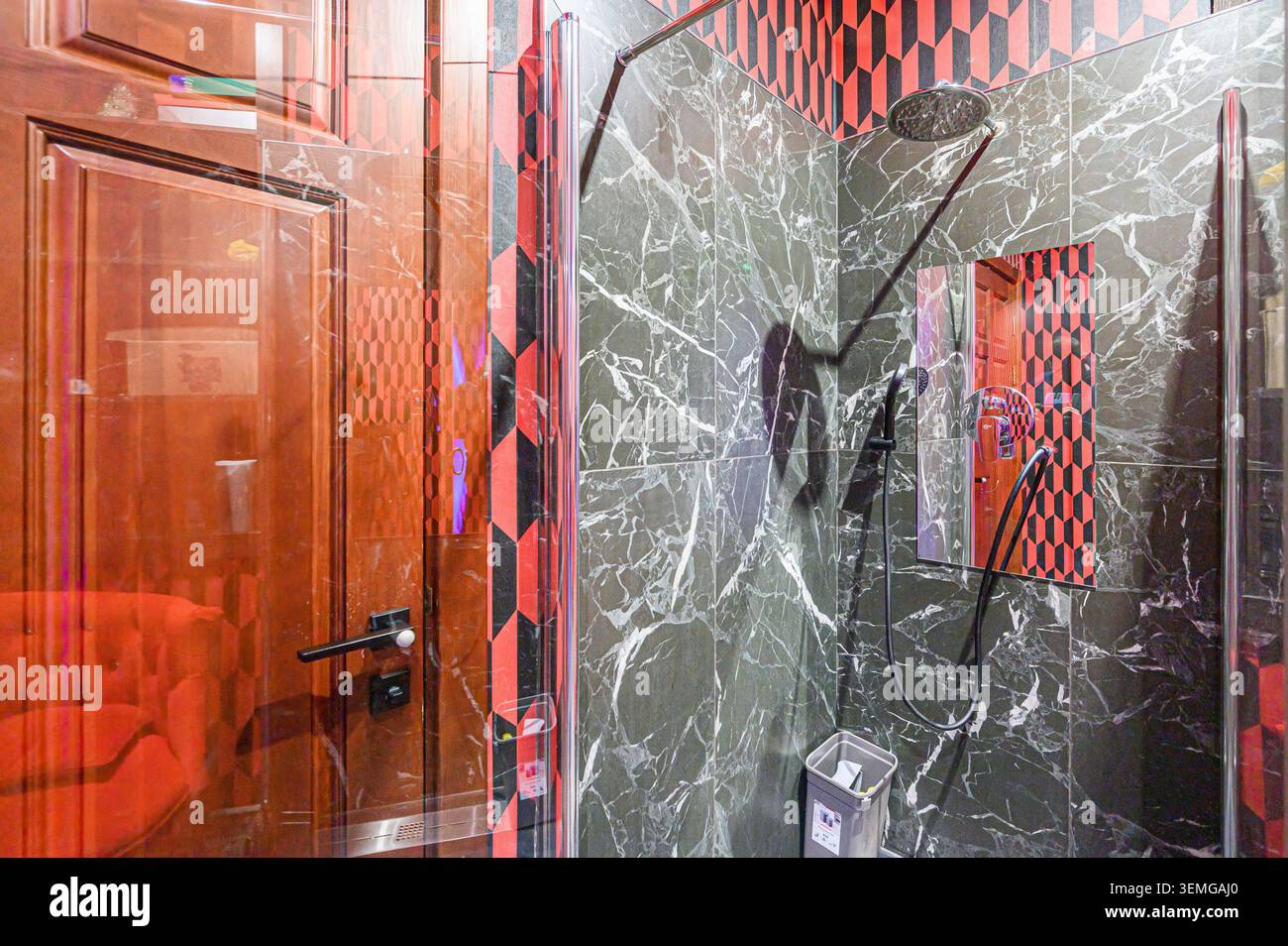 la stanza da bagno offre una cabina doccia con piastrelle in marmo, dettagli geometrici neri e rossi e una porta riflettente in legno. Foto Stock