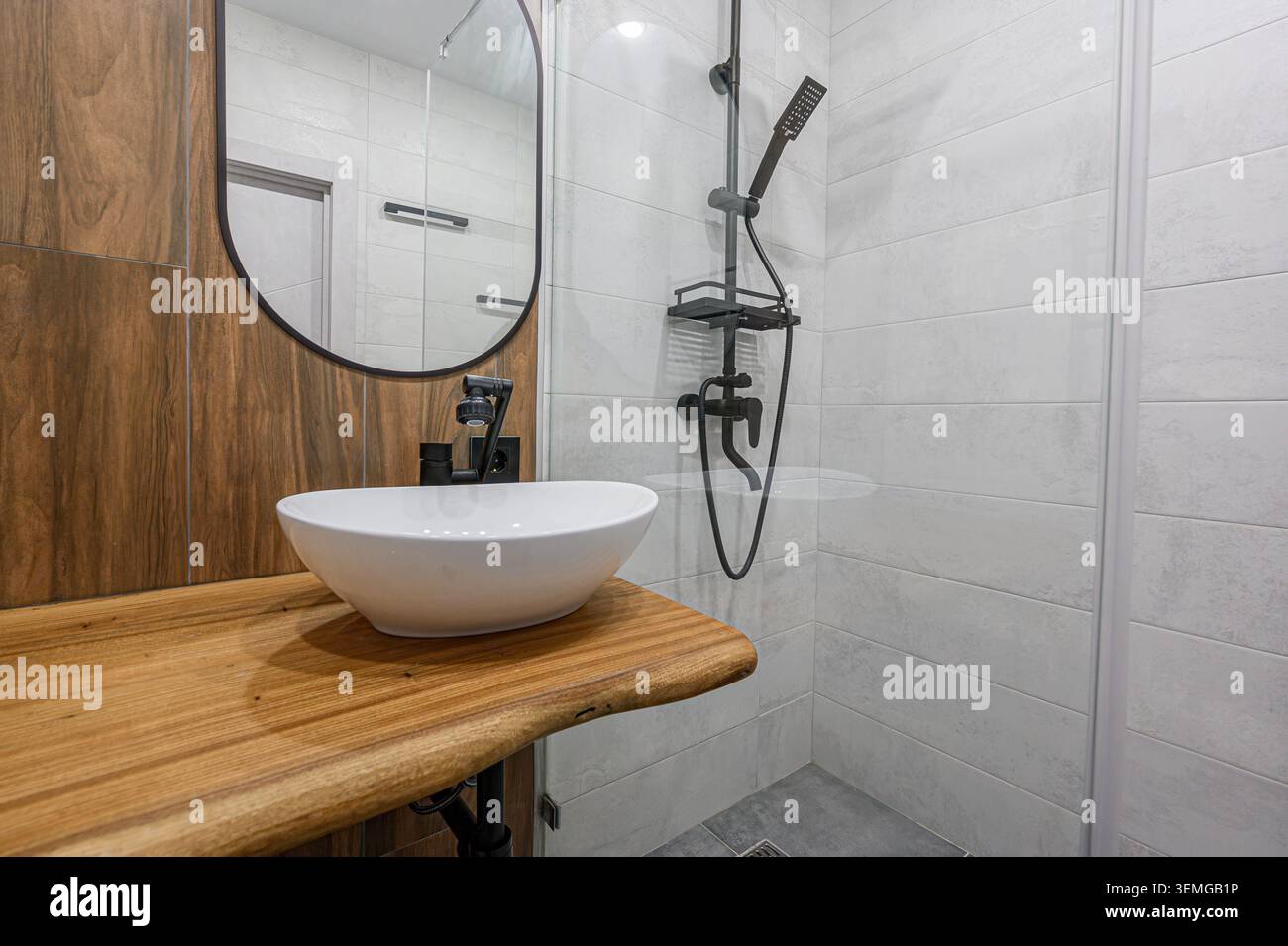 Bagno moderno con lavabo bianco su un ripiano in legno, accentuato da un rubinetto e uno specchio neri. Doccia con piastrelle bianche e finiture nere Foto Stock