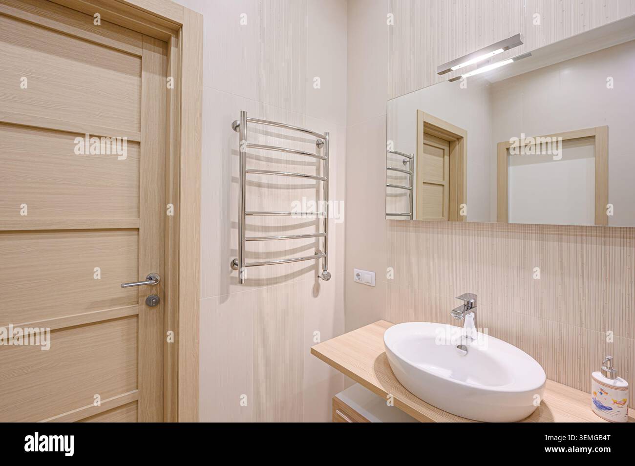 La vista del bagno è caratterizzata da una porta in legno chiaro, scalda asciugamani cromati, lavandino ovale sul ripiano in legno, specchio e piastrelle a righe neutre. Foto Stock