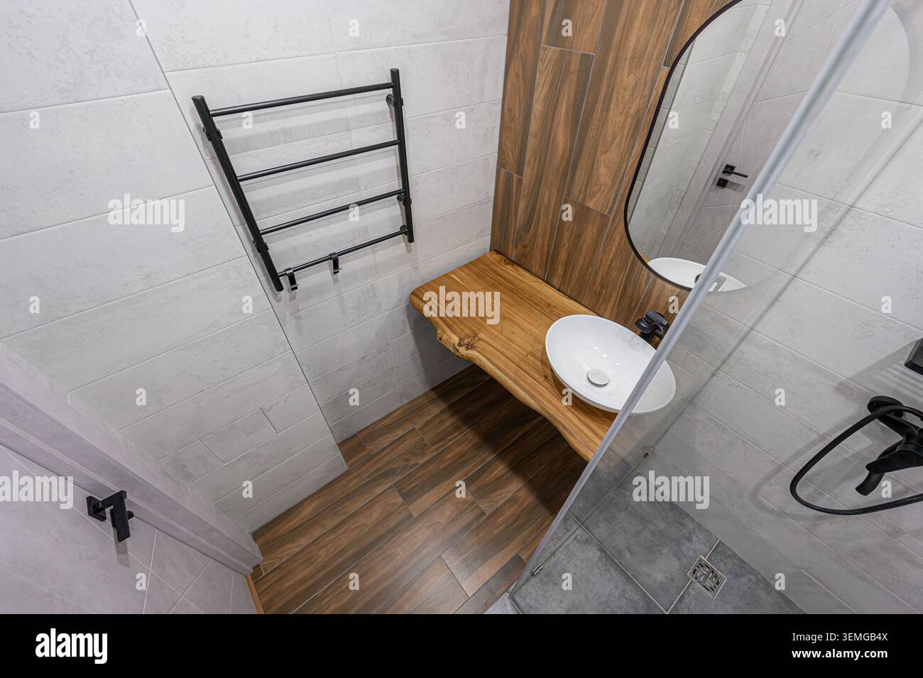 Bagno moderno con accenti in legno: Un lavandino con ripiano, specchio ovale, lavabo bianco e finiture nere. Piastrelle grigie e di legno Foto Stock