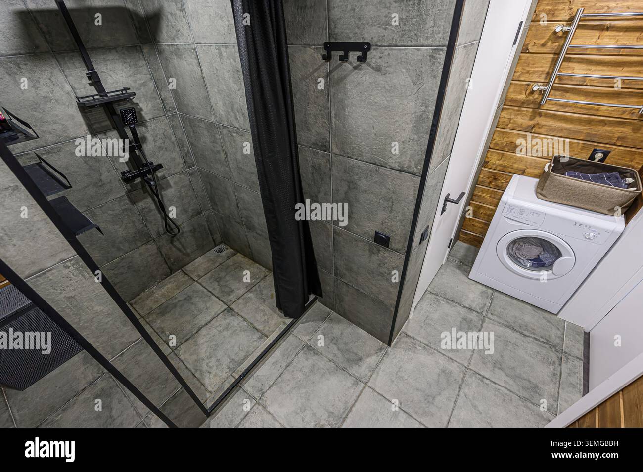bagno moderno con piastrelle grigie, doccia con cornice nera, lavatrice bianca e accenti in legno. Il design generale è pulito e funzionale. Foto Stock