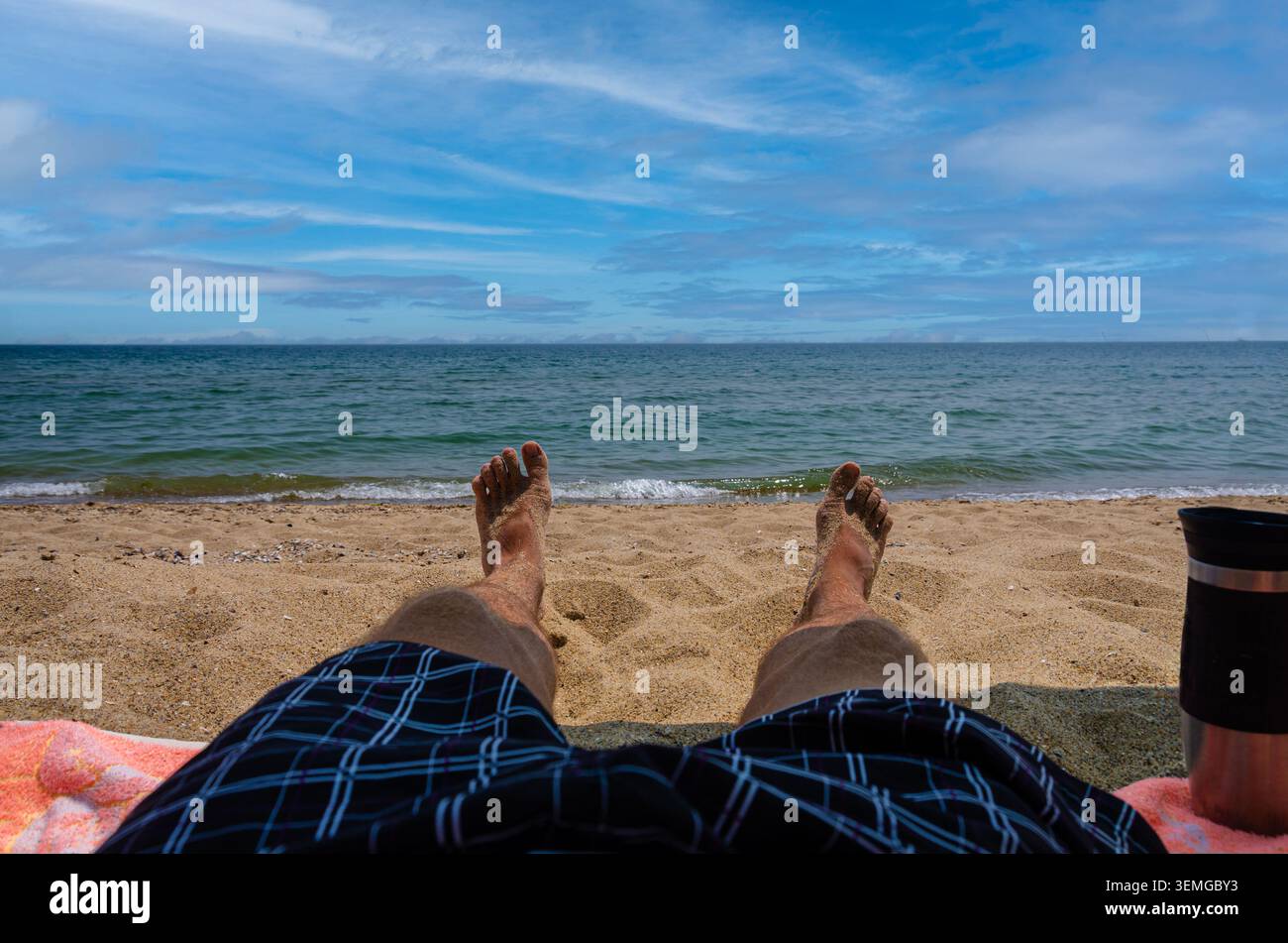 Una vista personale dei piedi e delle gambe di sabbia di un uomo in bauli a quadri, prendendo il sole su un asciugamano in spiaggia con vista sul mare blu calmo, Foto Stock