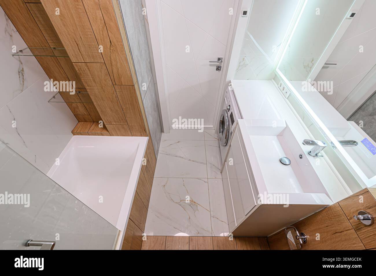 Bagno luminoso con vasca, pavimento in piastrelle di marmo bianco, un lavandino moderno e accenti in legno. Dispone di una lavatrice e di uno specchio illuminato Foto Stock