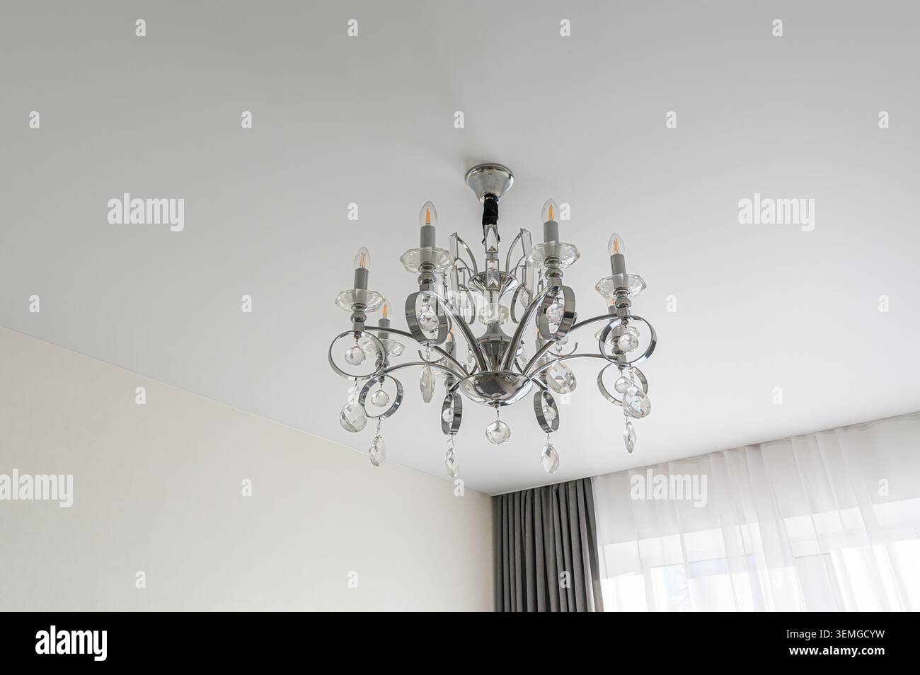 il lampadario di cristallo è appeso a un soffitto bianco, adornato da sei lampadine in stile candela e cristalli penzolanti. Le tende sono parzialmente visibili sul retro Foto Stock