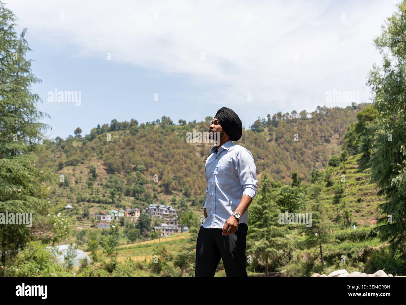 Un colpo ad angolo basso di un giovane Sikh che indossa un turbante nero e abiti formali che guarda verso il cielo mentre posa a Himachal Pradesh, India. Foto Stock