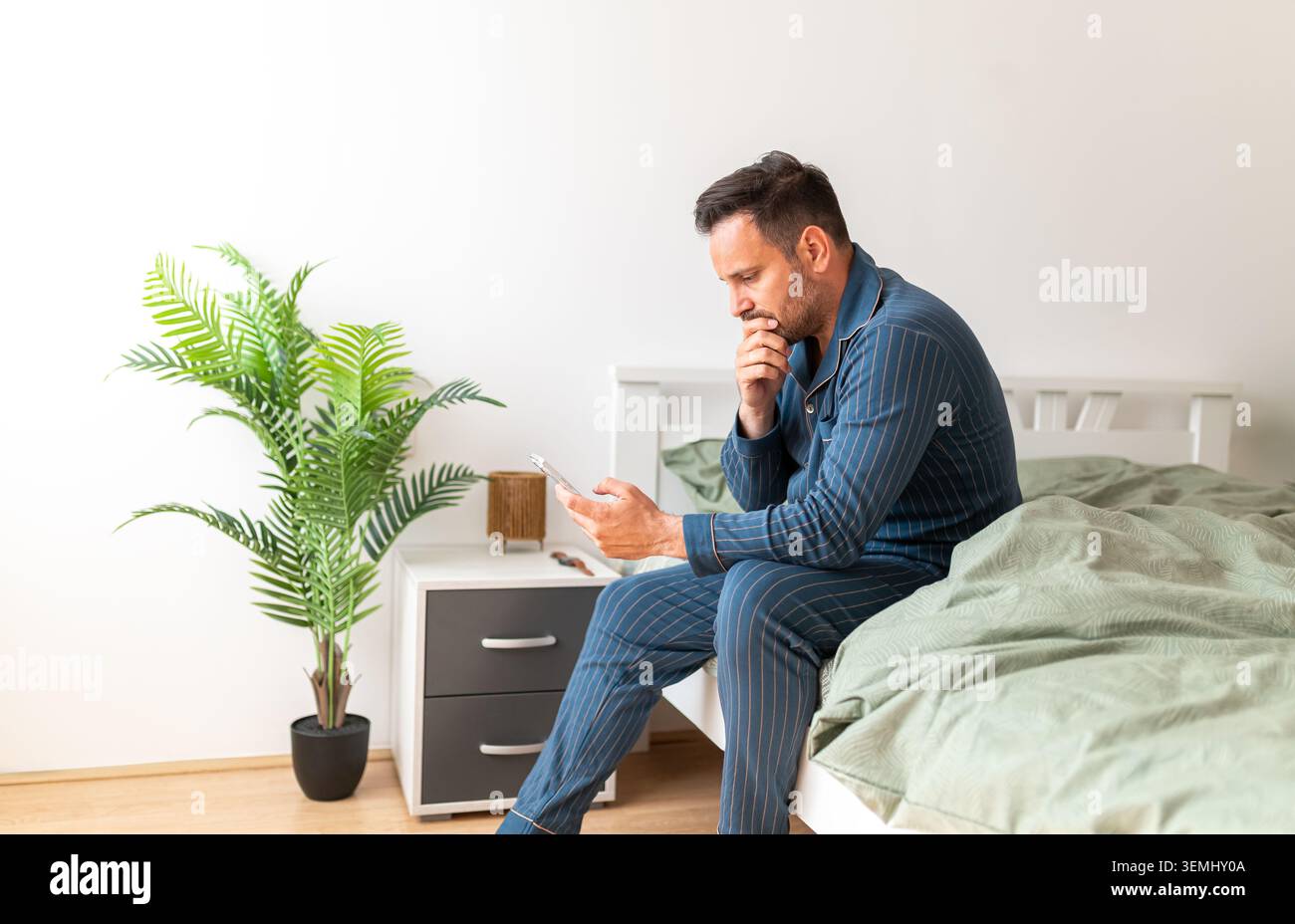 Uomo in pigiama seduto sul letto con attenzione, utilizzando uno smartphone, controllando le notizie o i messaggi del mattino, sperimentando stress o preoccupazioni, concentrandosi sulle cifre Foto Stock