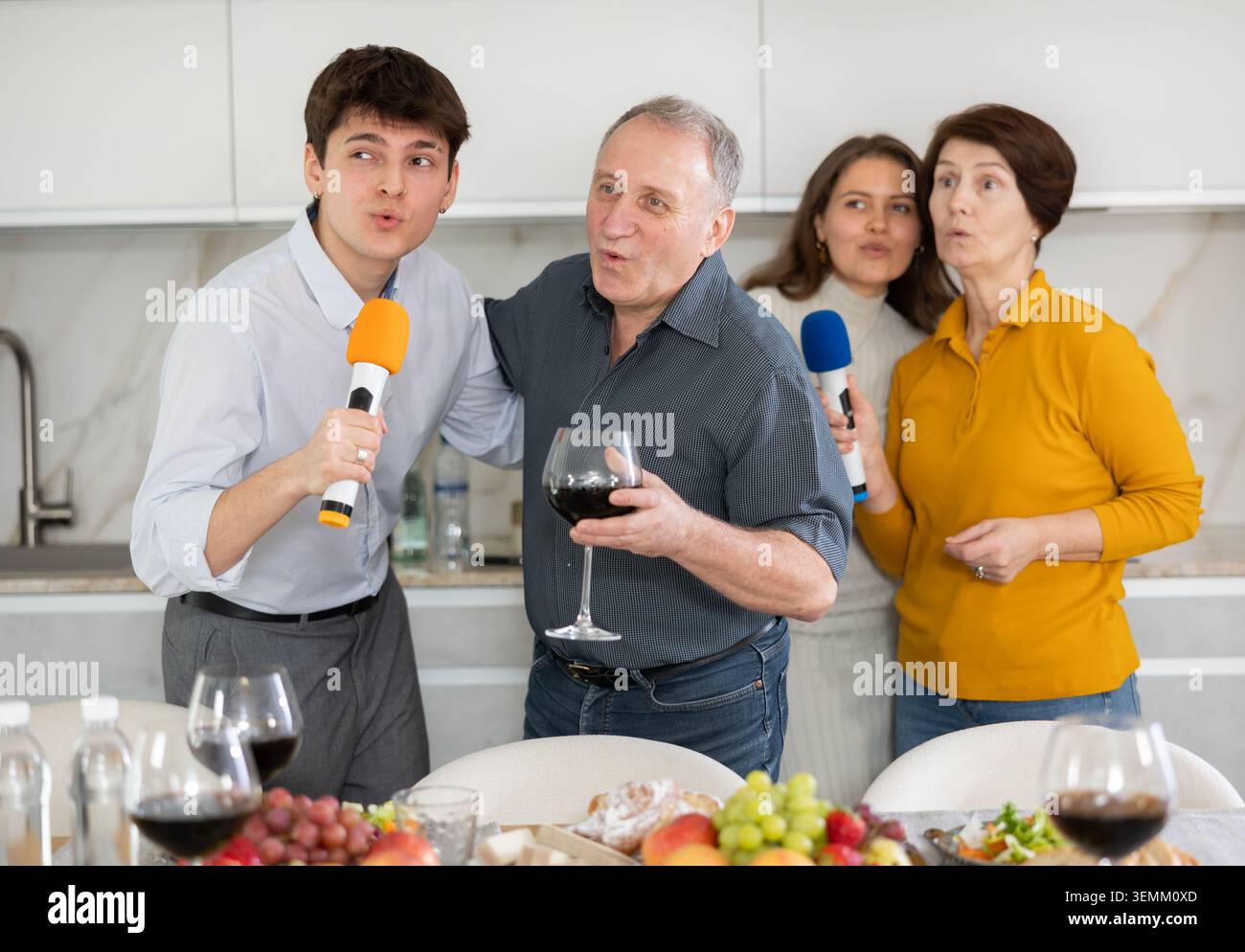 Giovani e genitori che si incontrano a casa si divertono al tavolo festivo in cucina, cantando eccitato karaoke Foto Stock